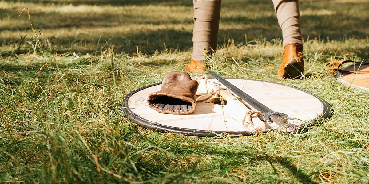 Foot wearing a tan shoe on a circular manhole cover in grass, with a person’s legs nearby
