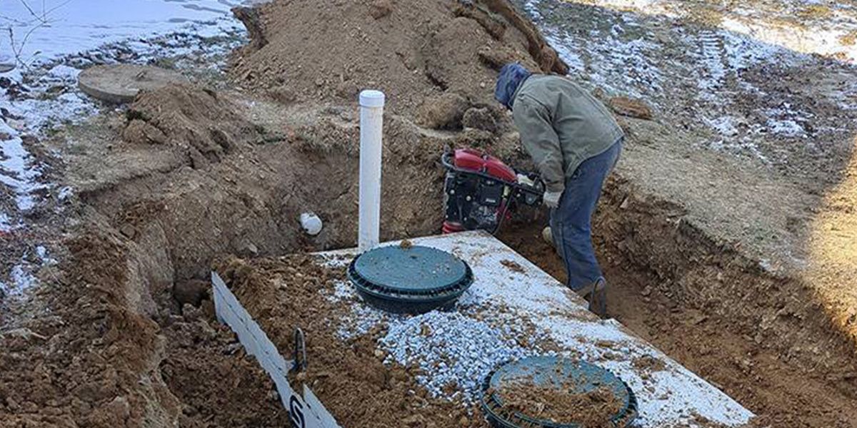 Worker digging near a septic tank in a muddy yard, with exposed pipes and covers.