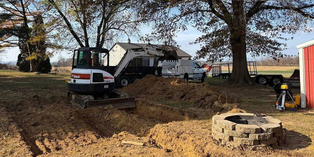 Excavator and skid steer working on a dirt lot beside a house with a stone fire pit in the foreground