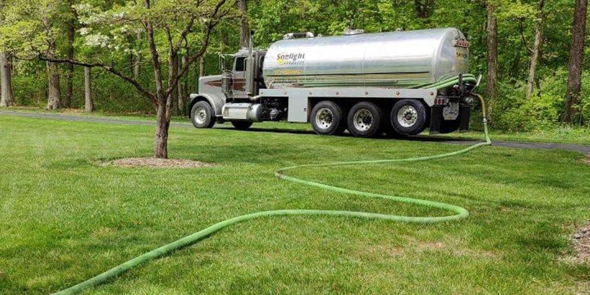 Silver tanker truck on grass near woods, with a green hose stretched across the lawn.