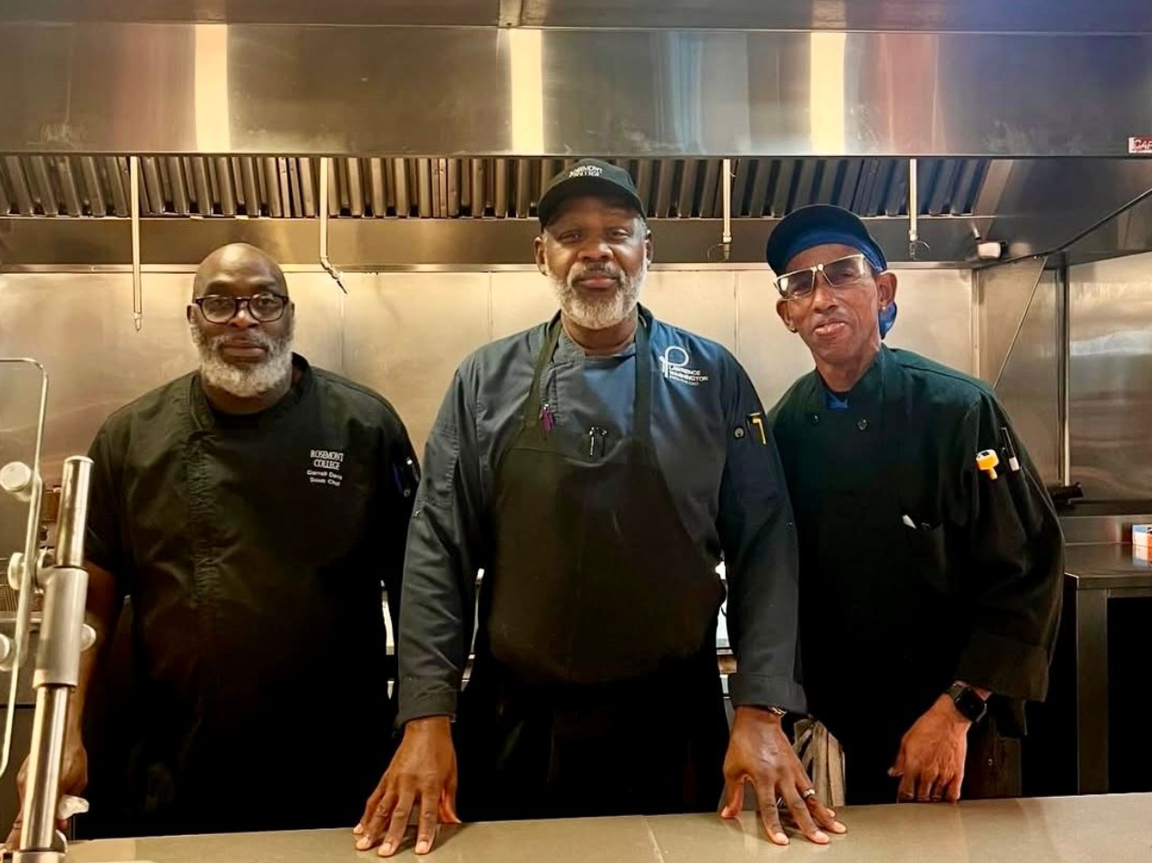 Three chefs standing in a commercial kitchen; stainless steel background.