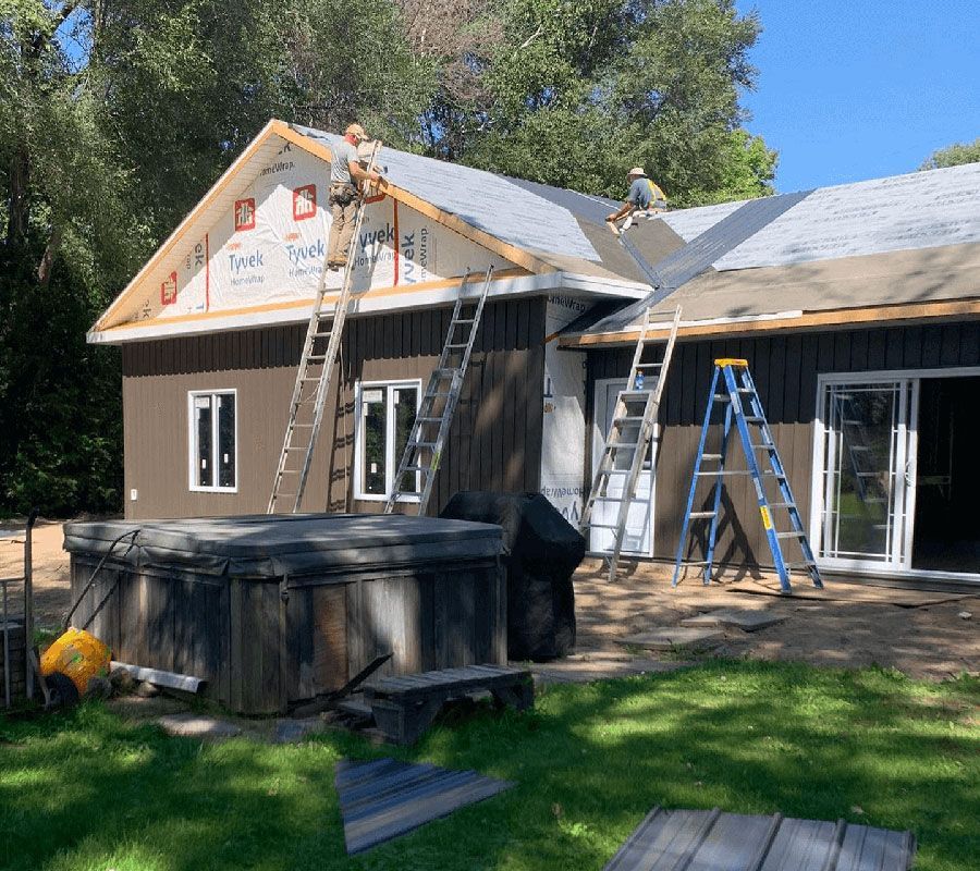 Two workers install roofing on a house exterior with ladders, a hot tub, and trees in the background.