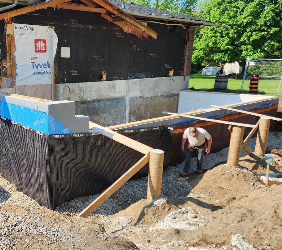 A construction worker stands by a building foundation featuring black drainage wrap, Tyvek sheeting, and wood bracing.