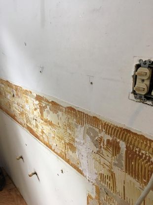 Damaged wall with exposed adhesive and removed tile backsplash beside an electrical outlet.