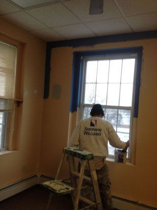 Person painting trim around a window in a room with peach walls and blue painter’s tape
