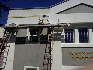 Worker on scaffolding painting the upper wall of a white building with gray trim.