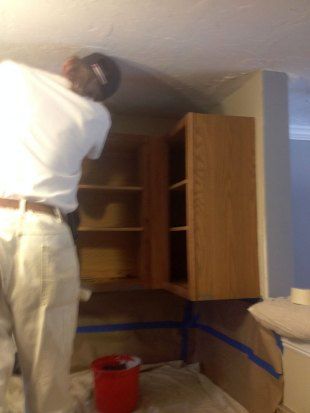 Person installing or adjusting wooden kitchen cabinets under a slanted ceiling, with blue painter's tape and a red bucket nearby.