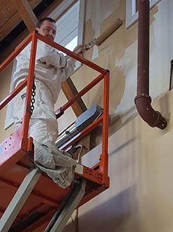 Worker in white coveralls standing on a red lift platform next to a wall-mounted pipe.