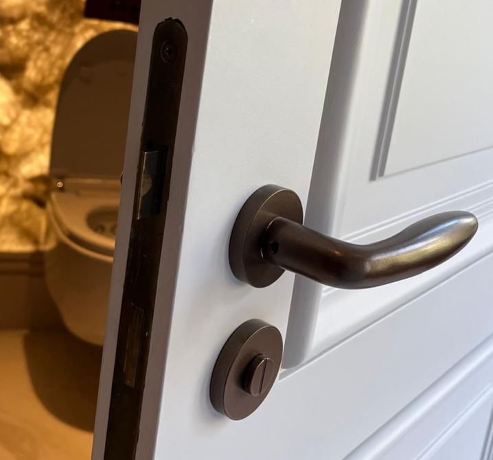 Koperen deurklink op een witte deur, met een warmgetinte kamer en een oranje stoel op de achtergrond.