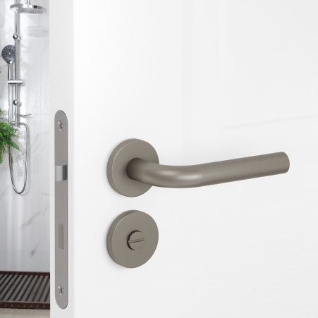 Close-up van een geborstelde metalen deurklink en slot op een witte badkamerdeur, met daarachter een douchecabine.