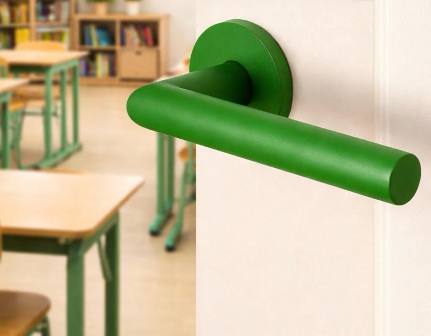 Groene horizontale deurklink op een witte klaslokaalmuur met bureaus op de achtergrond.
