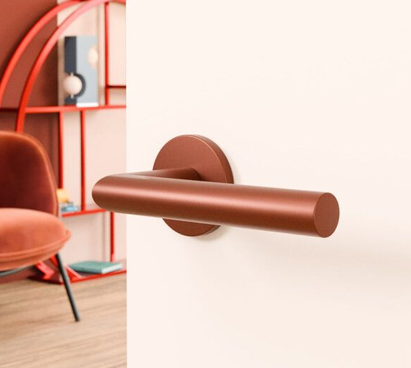 Koperen deurklink op een witte deur, met een warmgetinte kamer en een oranje stoel op de achtergrond.