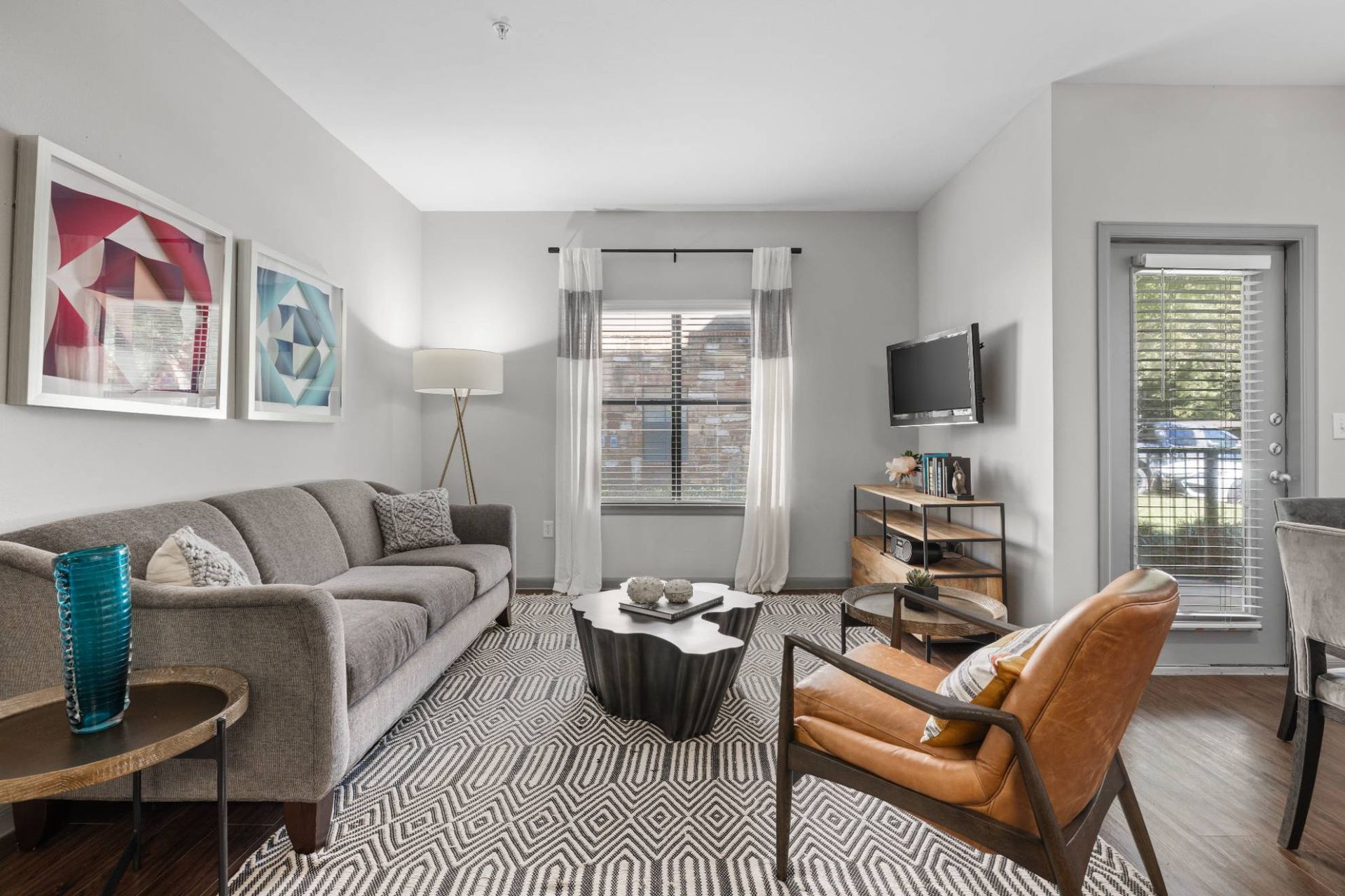 Living room of a modern apartment with a gray sofa, leather chair, geometric rug, and a wall-mounted TV.