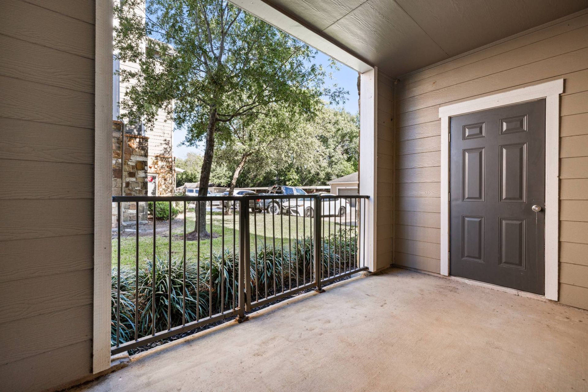 Covered apartment balcony with a gray door, black railing, and view of trees and parked cars.