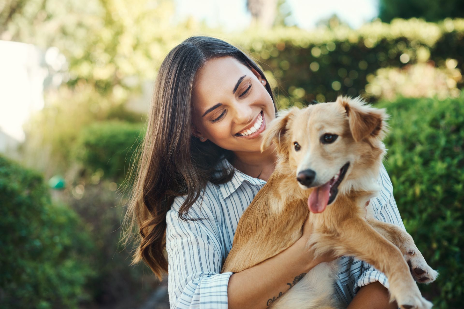 Woman smiling and holding a tan dog outdoors in a sunlit garden