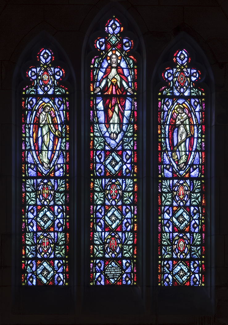Stained glass window in a church; central panel shows Jesus, flanked by figures in colorful, geometric patterns.