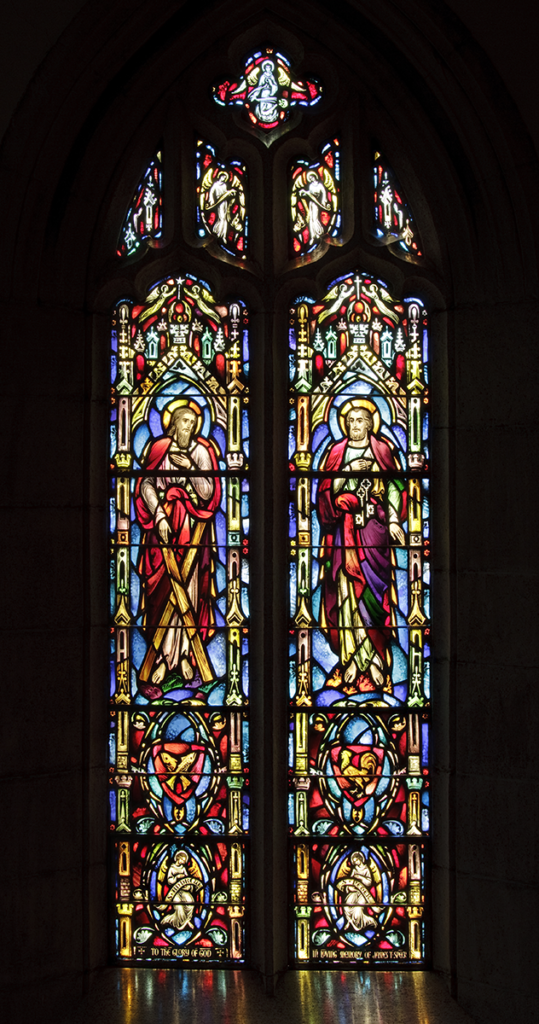 Stained glass window with two figures in robes; colorful and ornate, set in a dark arch.
