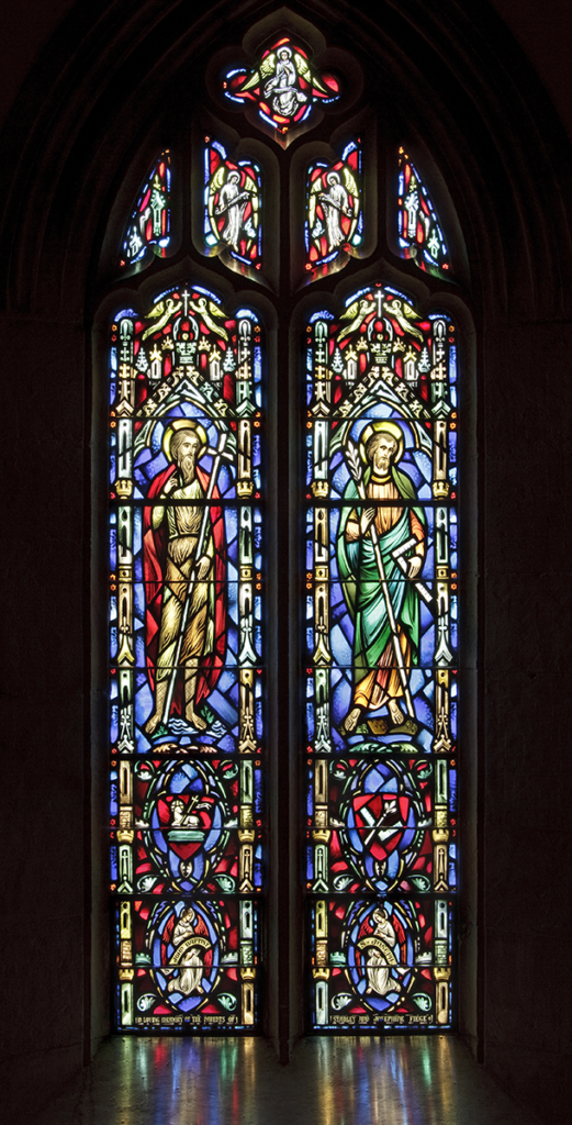 Stained-glass window with two saints in a church. Figures in red and green robes, surrounded by blue and colorful glass.