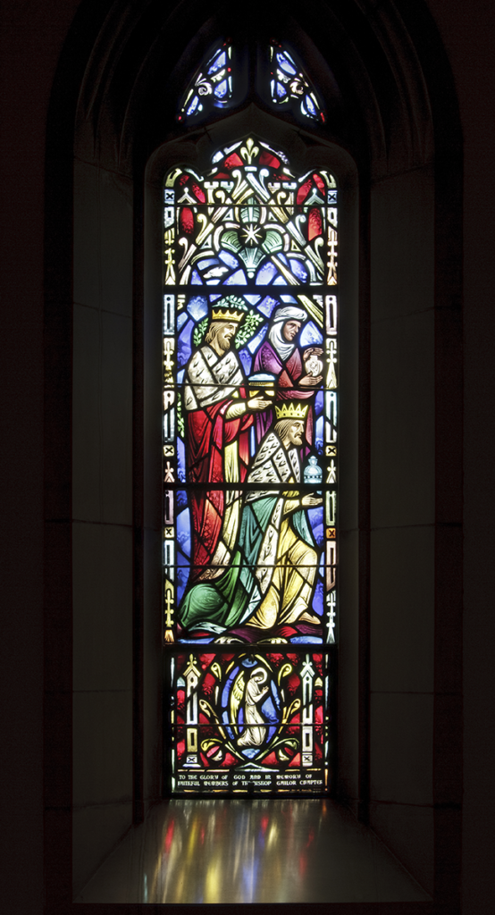Stained glass window depicts biblical figures: a king offering gifts to a baby, vibrant colors in a gothic setting.