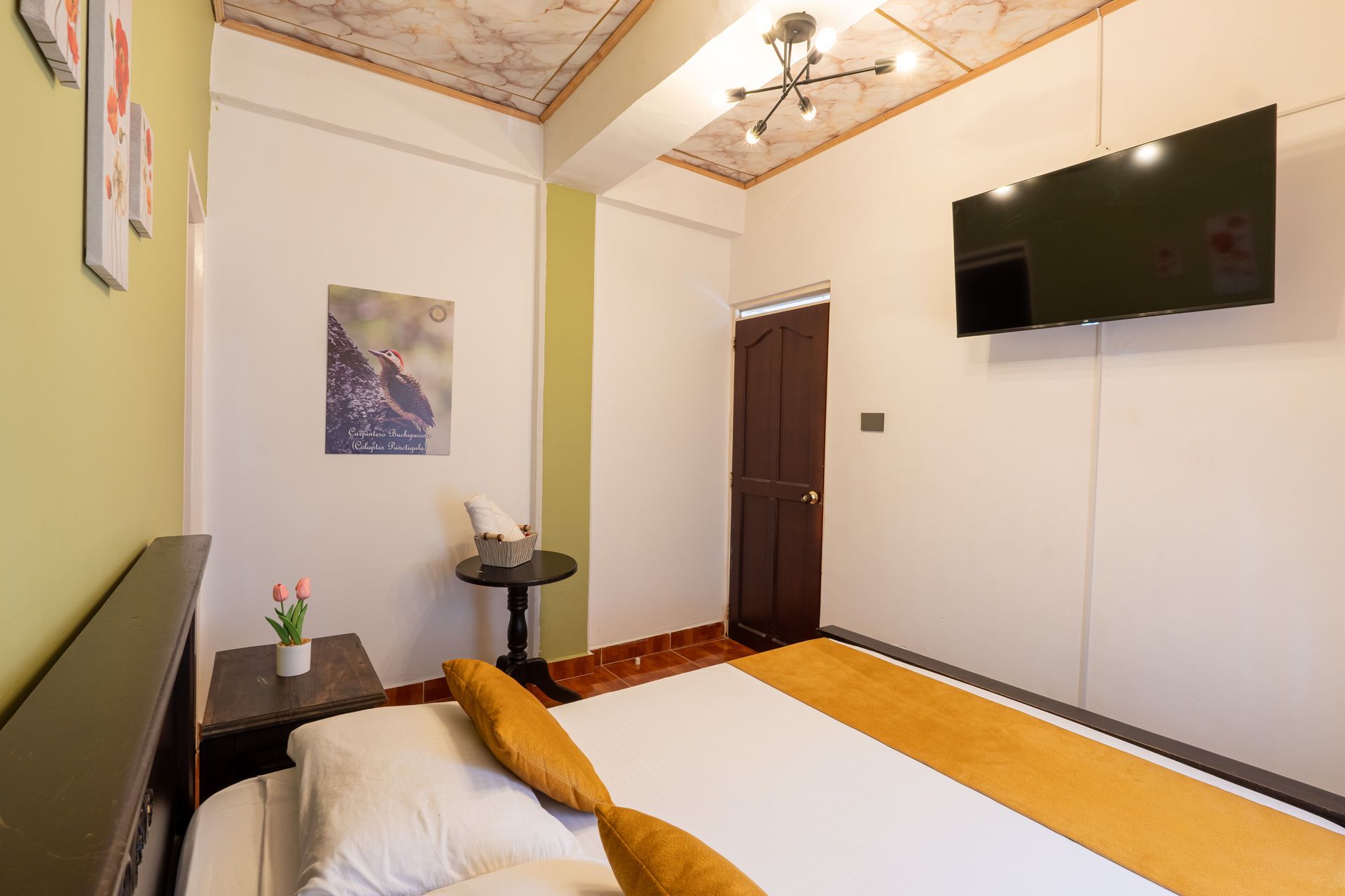 Habitación pequeña con cama, televisor montado en la pared, puerta oscura y decoración cálida en naranja y blanco.