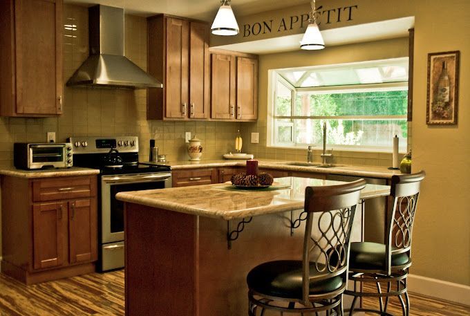 A warm-toned kitchen with wood cabinets, a stainless steel range, marble-topped island with two chairs, and a window.