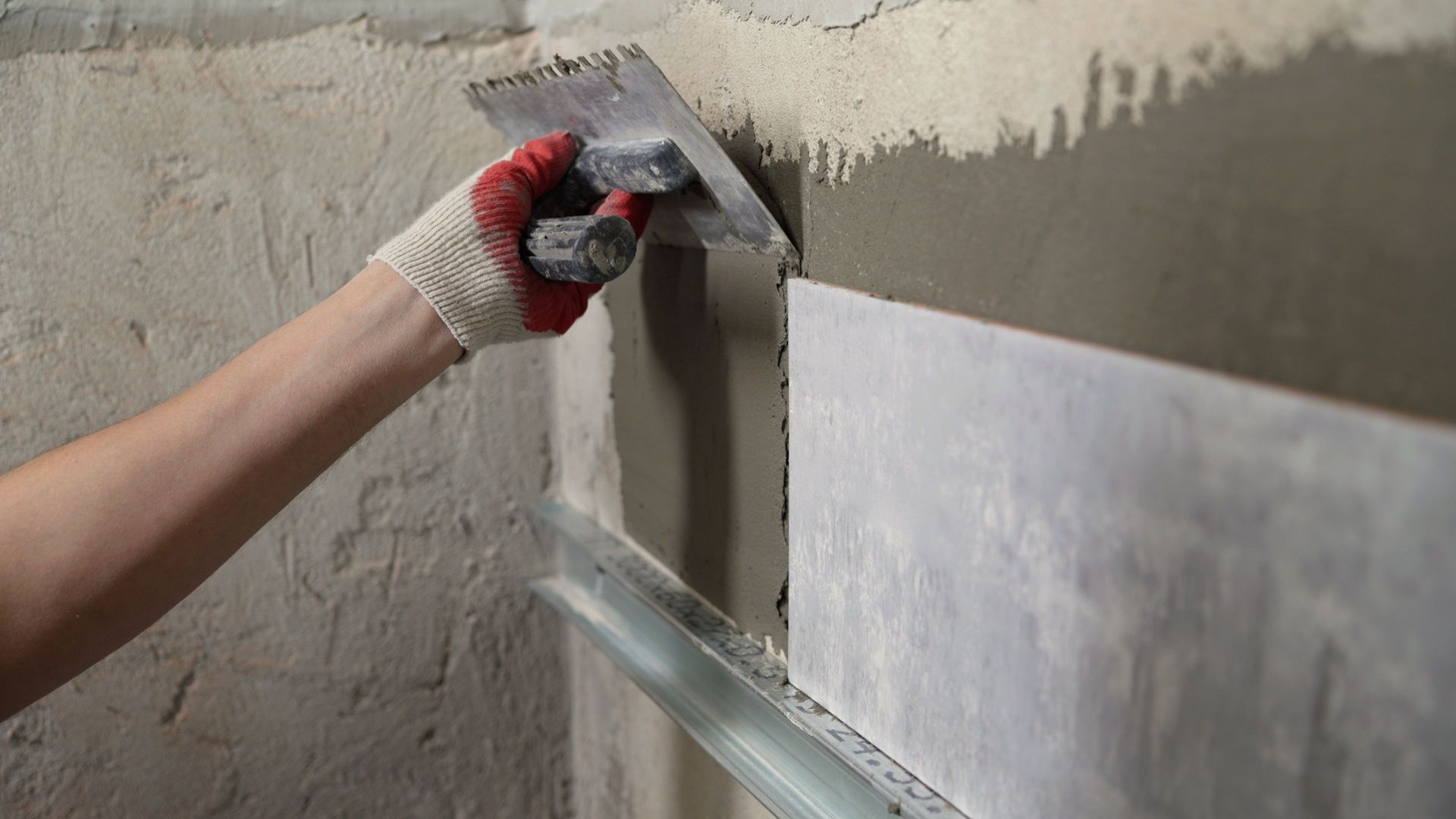 A worker wearing a red and white glove uses a notched trowel to spread mortar on a wall before applying a tile.