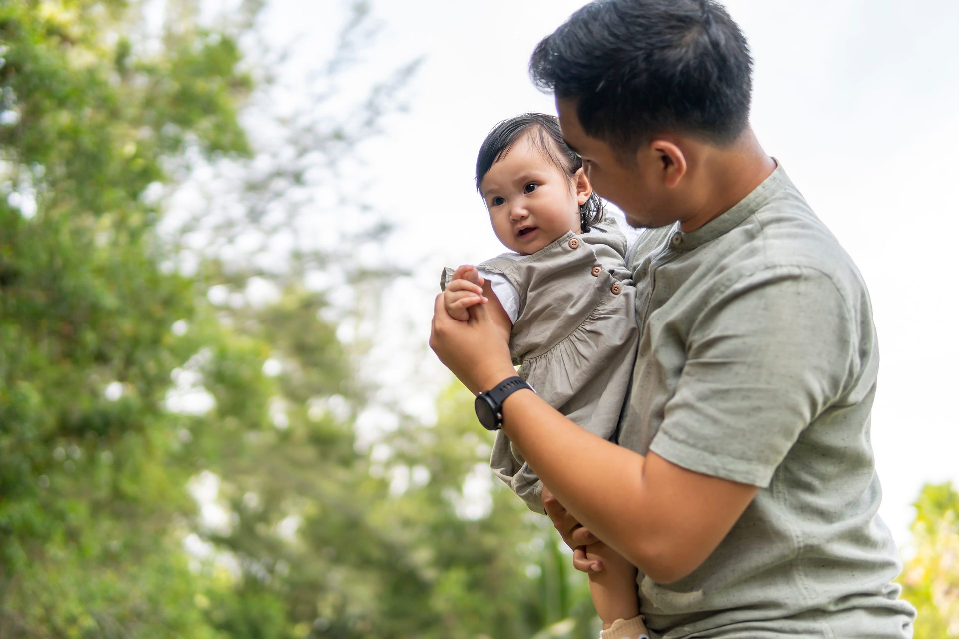 Adult holding smiling baby outdoors in a park with green trees