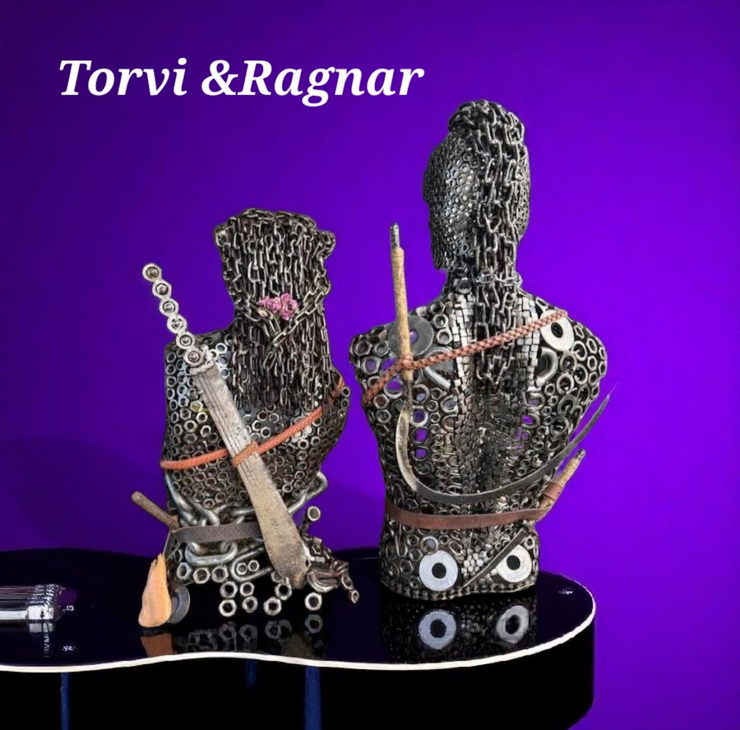 Metal sculptures of two figures on a shiny black surface, against a purple backdrop. Text reads