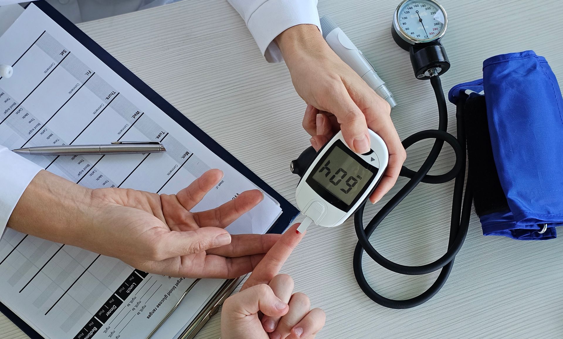 Person testing blood glucose with a meter; stethoscope and medical forms are nearby. Person testing blood glucose with a meter; stethoscope and medical forms are nearby.