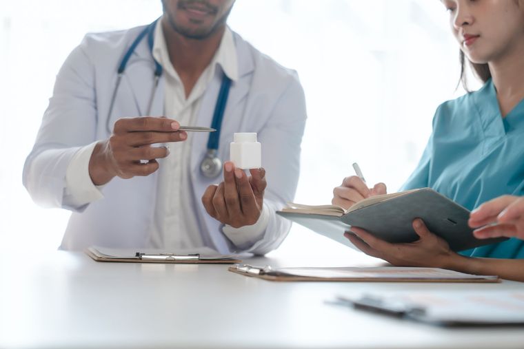 Doctor showing medication bottle to a person in scrubs writing notes at a table.