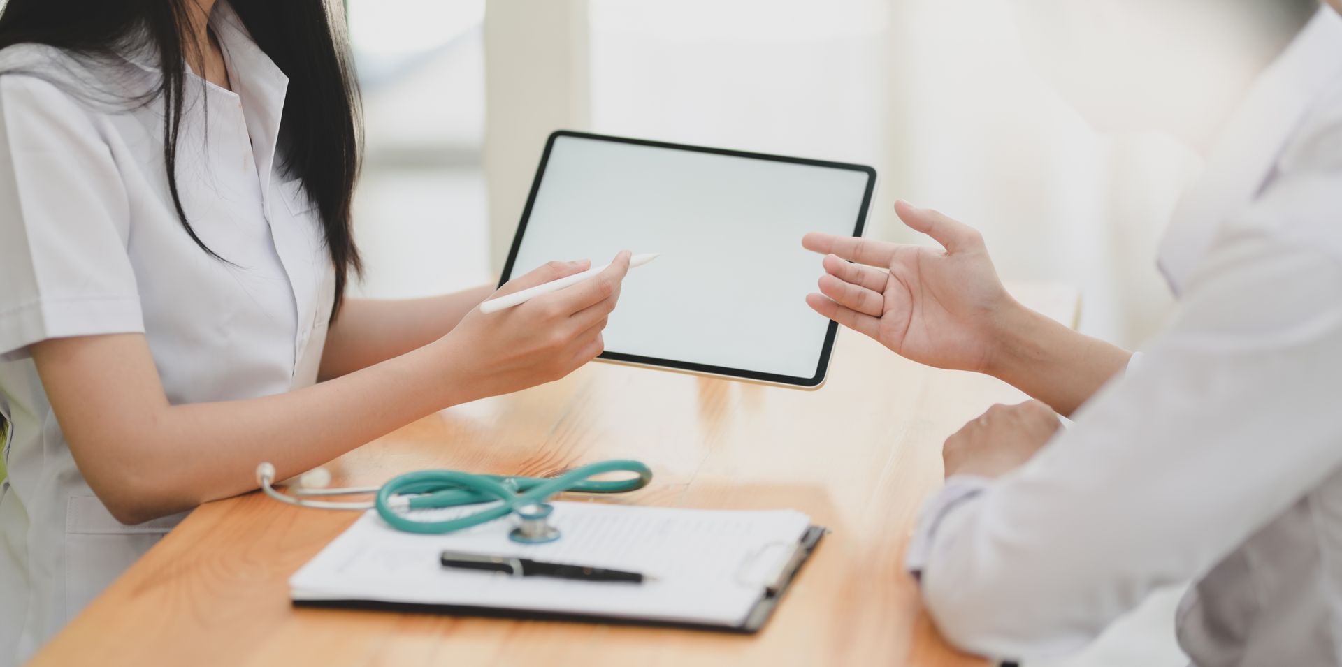 A medical professional points at a tablet screen while consulting with a patient at a desk. A medical professional points at a tablet screen while consulting with a patient at a desk.