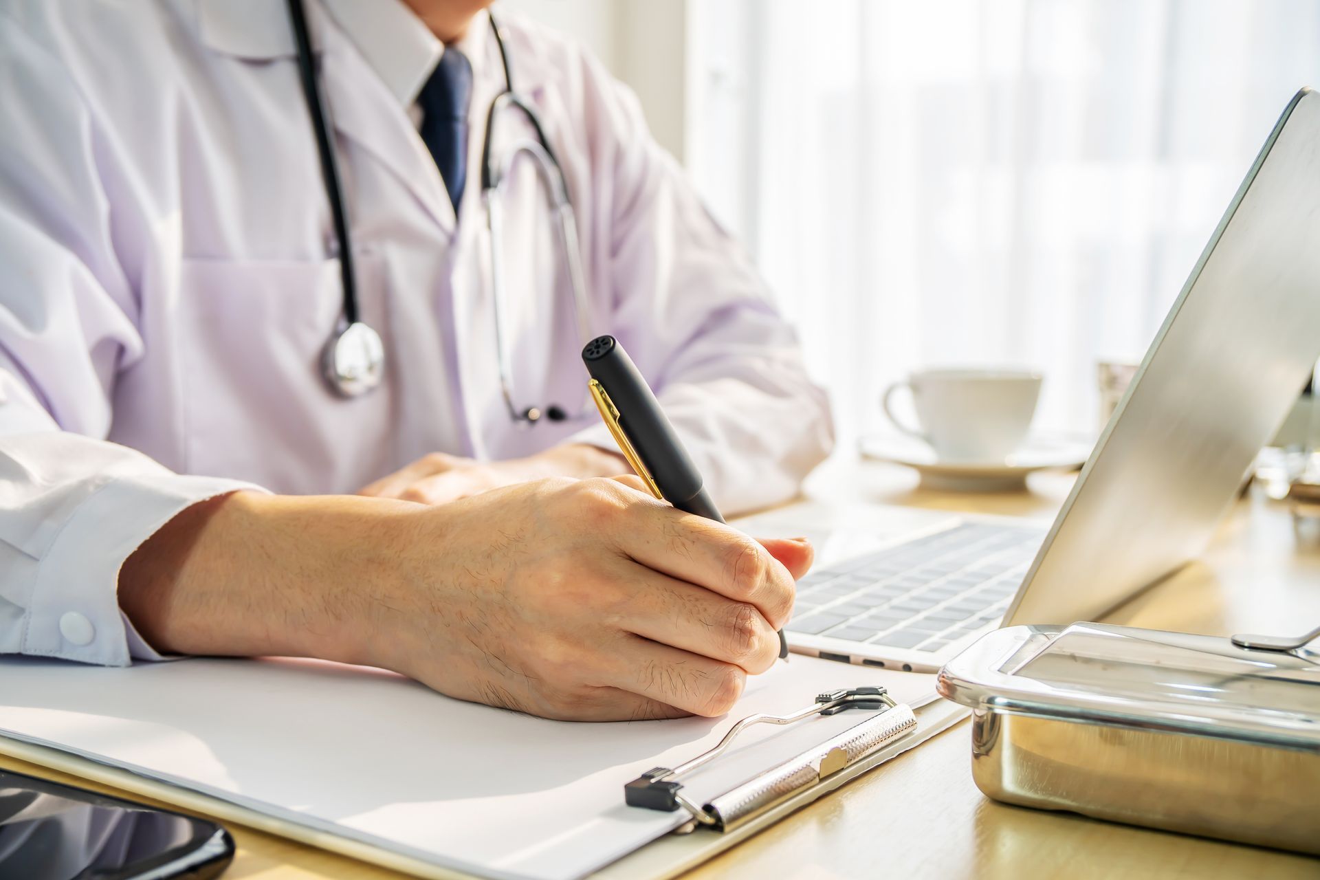 Doctor writing on a clipboard at a desk, laptop and stethoscope present.
