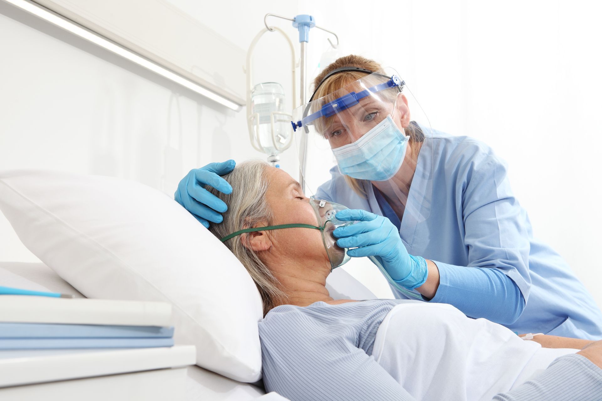 Nurse in blue scrubs, mask, and face shield assists a patient with an oxygen mask in a hospital bed. Nurse in blue scrubs, mask, and face shield assists a patient with an oxygen mask in a hospital bed.