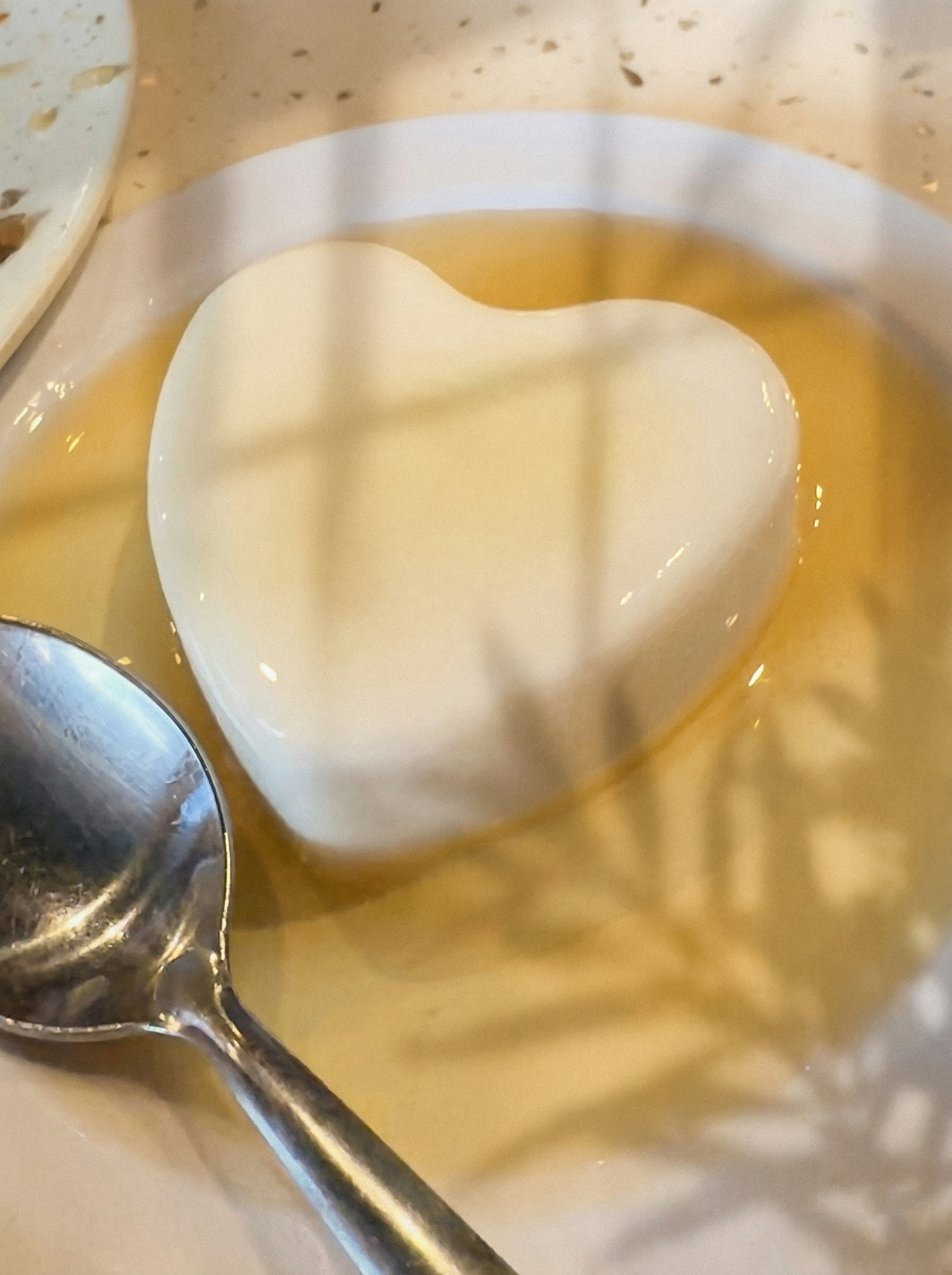 A heart-shaped white dessert sits in a pool of golden syrup on a white plate, with a silver spoon to the side.