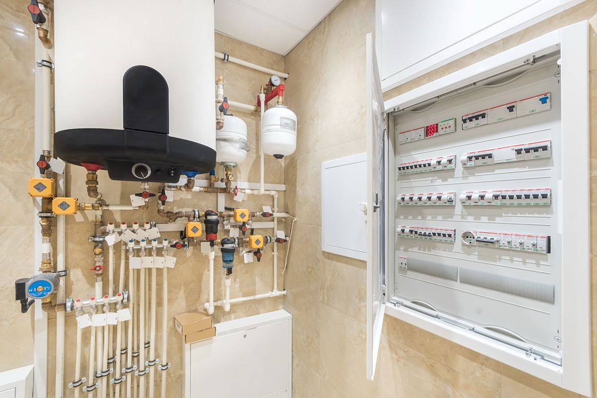 Plumbing and electrical systems in a utility room. White pipes, water tanks, and circuit breakers against beige tiled wall.