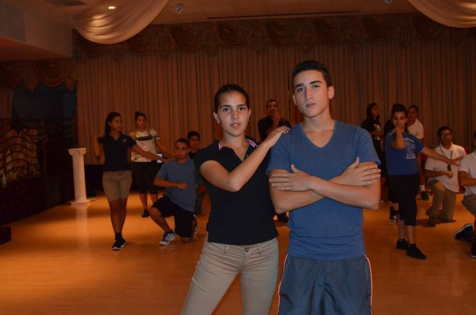 Two people posing with arms crossed on a dance floor, with other dancers in the background.