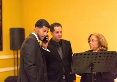 Three people in suits converse beside a music stand in a warm indoor room.