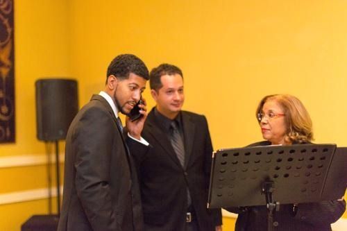 Three people in suits stand at a microphone, speaking in a yellow-walled room.