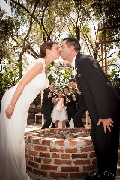 Bride and groom kissing beside a brick well under trees, with wedding guests watching nearby
