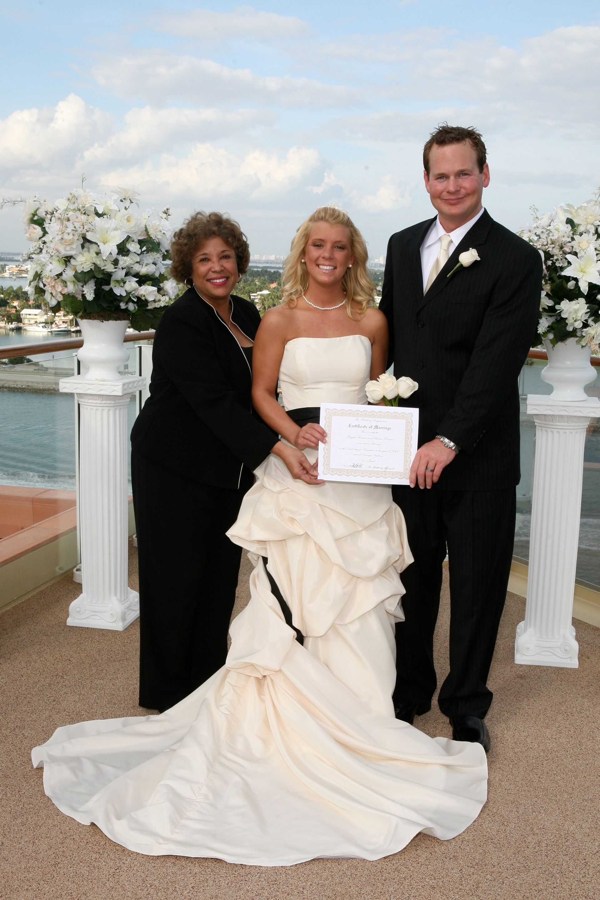 Bride and groom with a woman holding a certificate at an outdoor wedding by the water