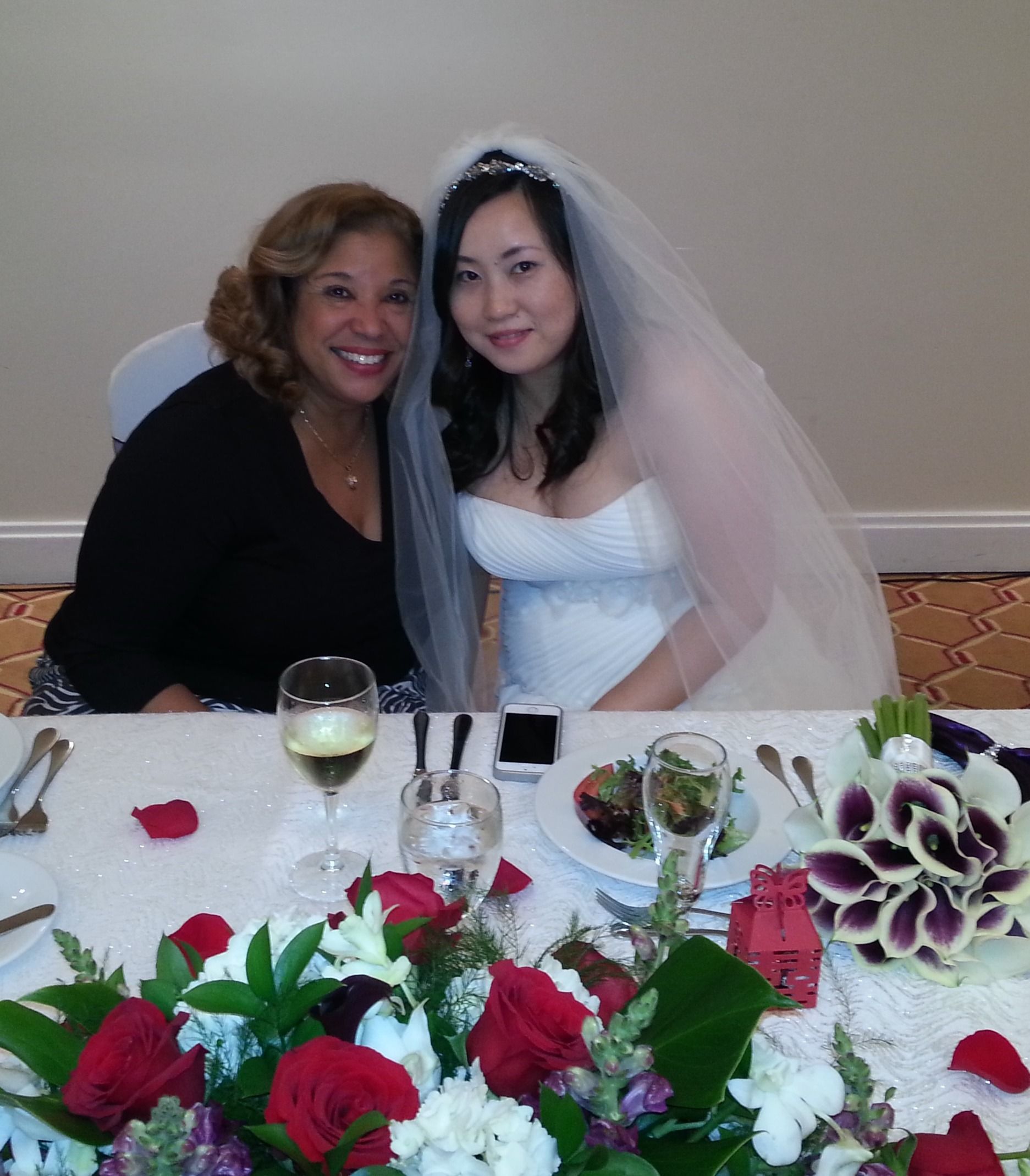 Bride and guest seated at a decorated wedding reception table, smiling beside floral centerpieces.
