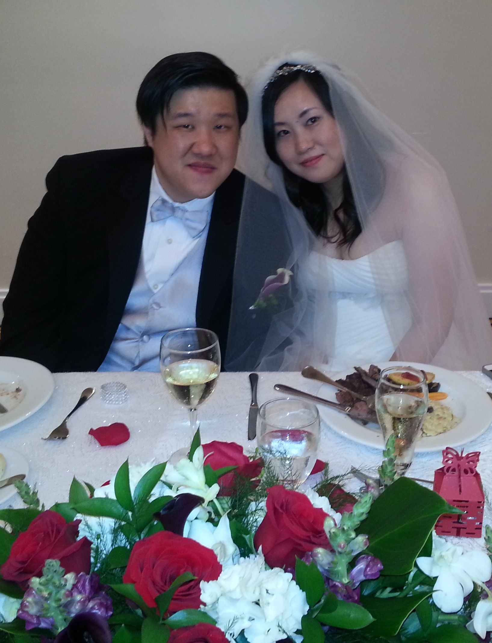 Bride and groom smiling at a wedding table with red and white flowers in the foreground