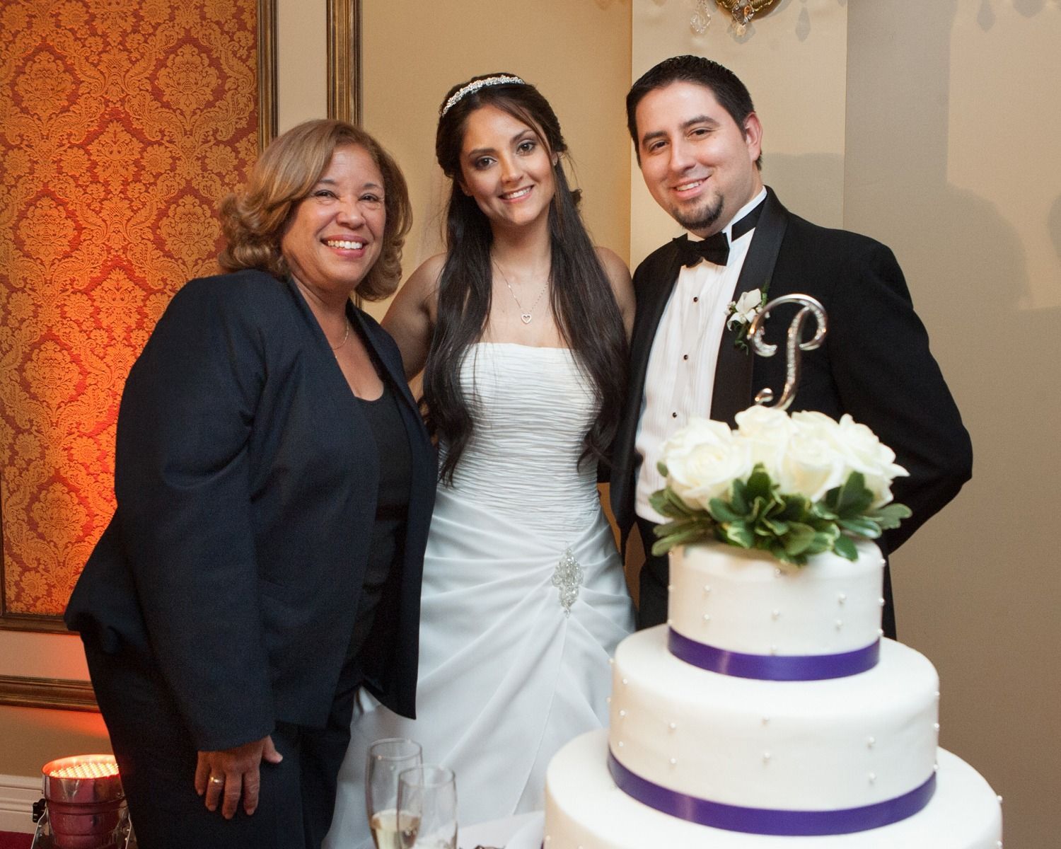 Three people posing beside a white wedding cake in a warmly lit banquet hall
