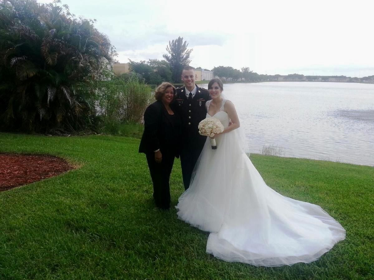 Three people posing on a grassy lakeside, with a bride in a white wedding dress holding a bouquet.