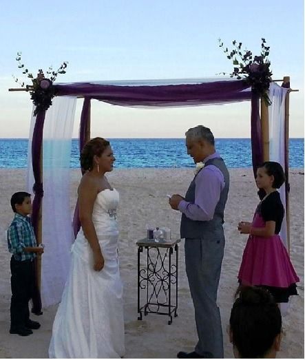 Beach wedding ceremony under a purple-draped arch by the ocean, with couple and two children nearby