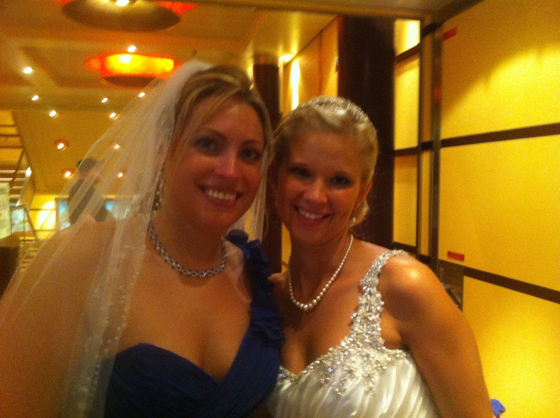 Two women in formal dresses smiling in a warmly lit hallway at an event