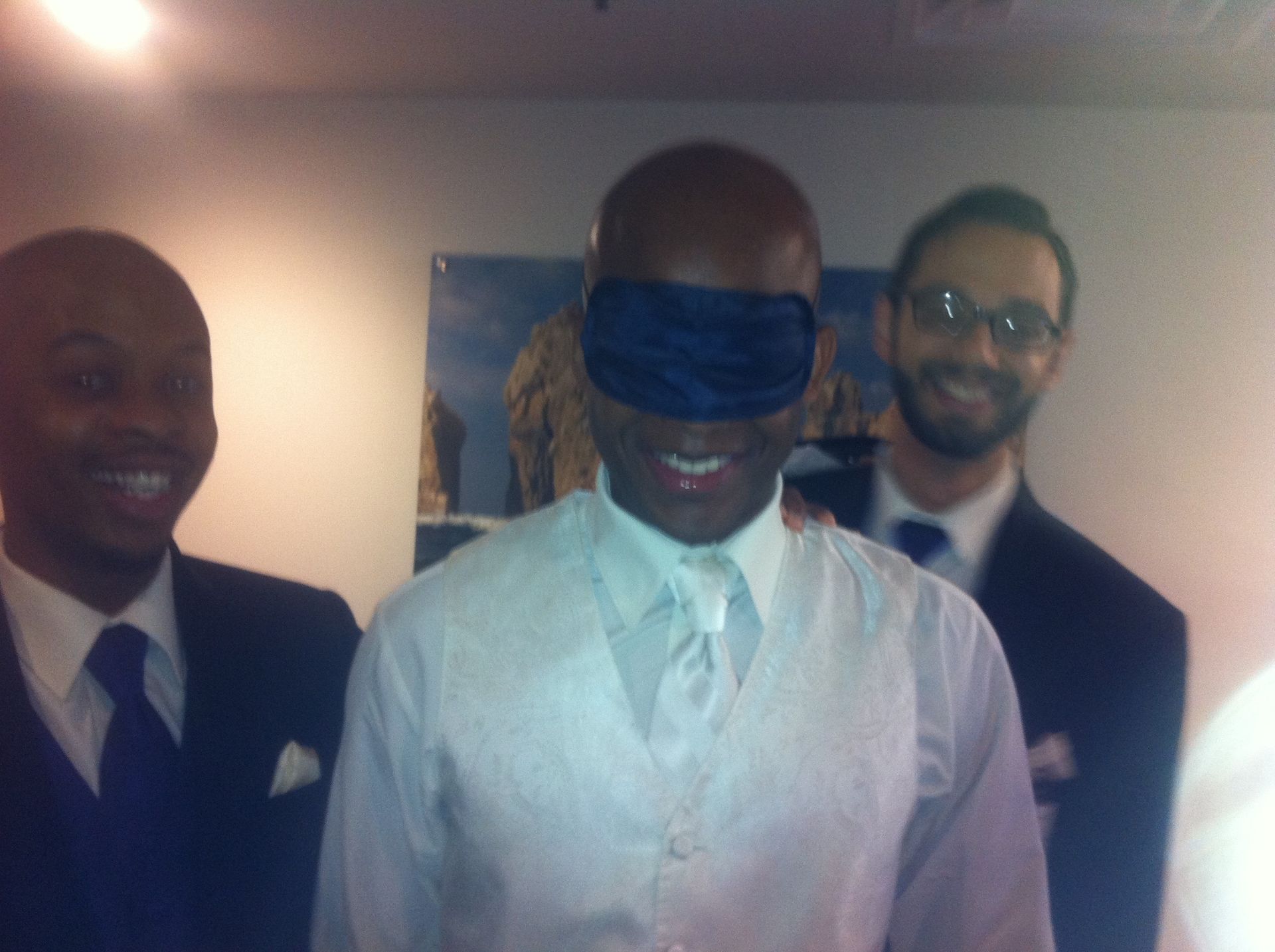 Three men in suits smiling indoors; the center man is blindfolded with a blue cloth.