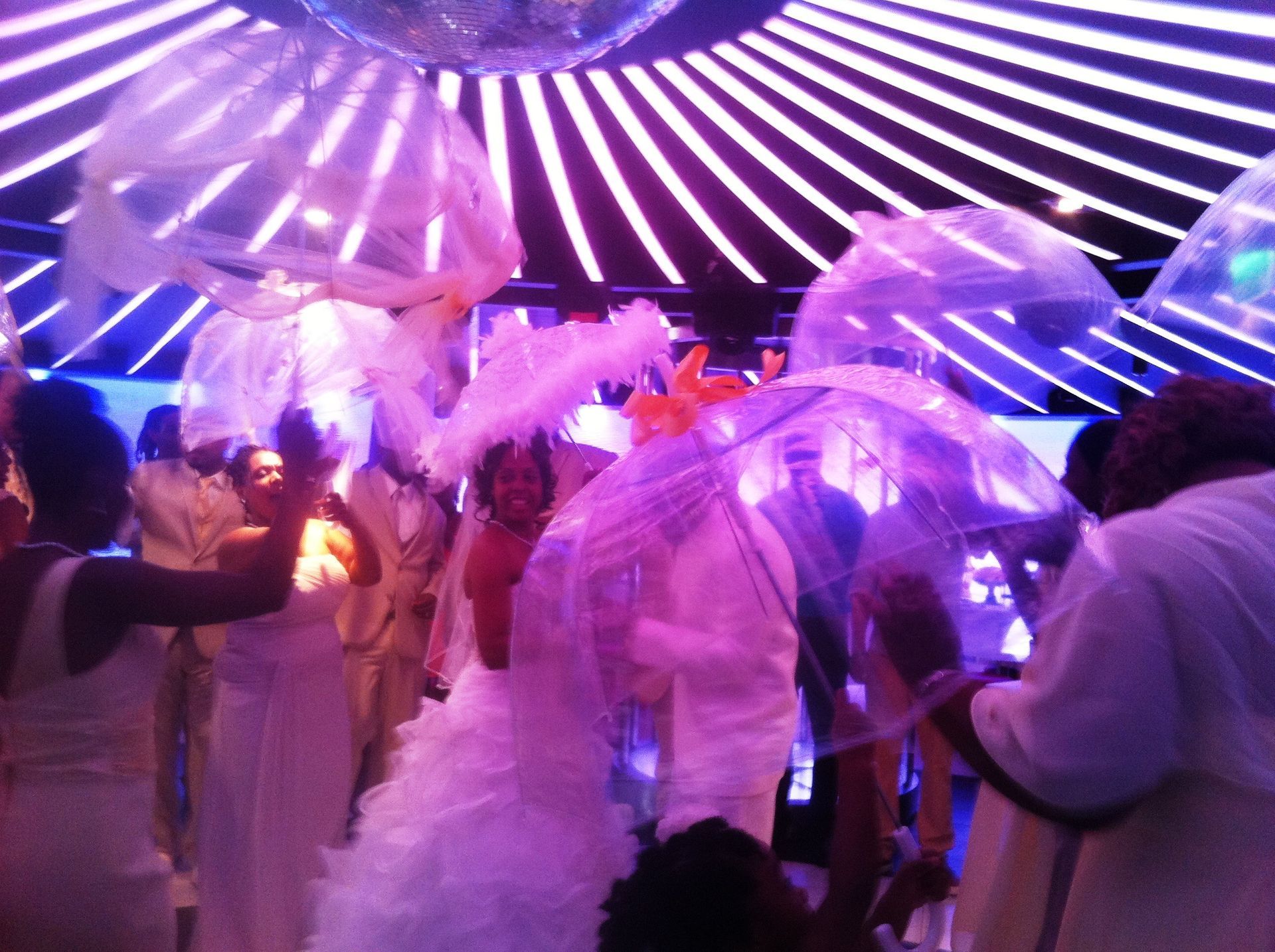 People dancing under purple and white lights at an indoor party, holding translucent umbrellas