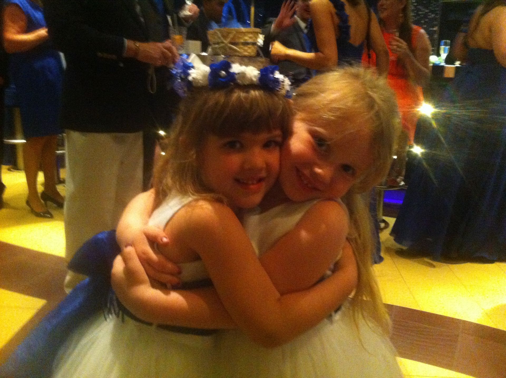 Two children in dresses hugging and smiling at an indoor event, one with a blue flower crown.