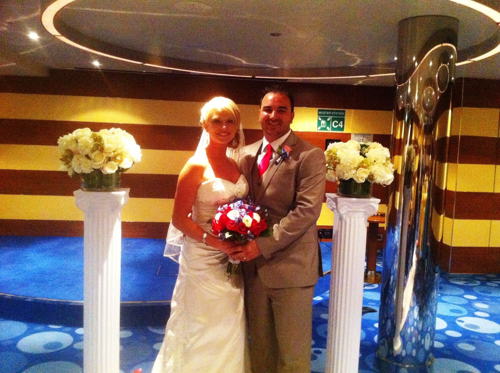 Bride and groom posing in a cruise ship lobby with a red-and-white bouquet and white floral arrangements
